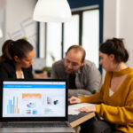Computador laptop aberto em primeiro plano, com 3 pessoas ao fundo analisando gráficos diversos