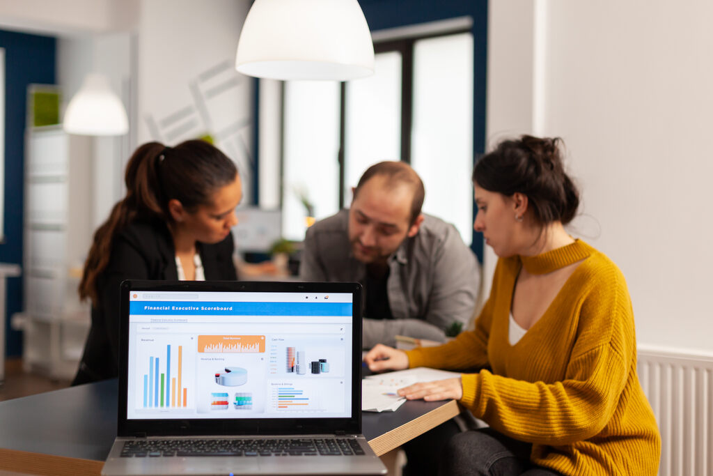 Computador laptop aberto em primeiro plano, com 3 pessoas ao fundo analisando gráficos diversos
