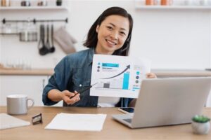 Mulher asiática jovem, de cabelos curtos, segurando um gráfico enquanto sorri em frente a um computador