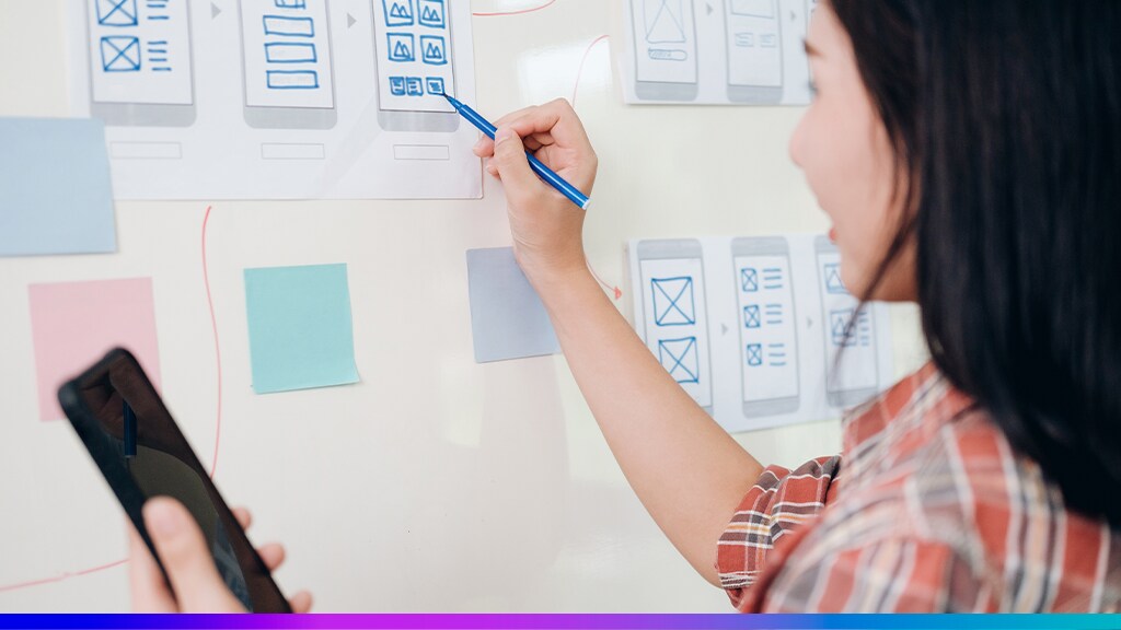 Profissional desenhando protótipo de interface mobile em um quadro branco com anotações e post-its, representando o processo de design centrado no usuário.