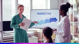 Profissionais apresentando planejamento estratégico de marketing em uma reunião, com gráfico em tela mostrando etapas de pesquisa, ideias e produção.